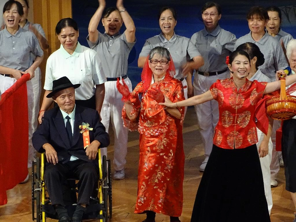 劉江南阿公(左)和劉尤碧真阿嬤(中)一起慶祝結婚五十年，志工巧扮媒婆(右一)帶著舞臺上、下，隨著樂曲打拍，全場充滿喜悅。[攝影者：洪順發]