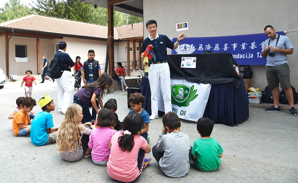 歐洲慈濟志工長期關心滯留在塞爾維亞的各國難民；慈濟志工發放新書包和文具用品給難民孩子。林正中師兄(惟揚，中)帶來布袋戲木偶劇，透過活潑可愛的表演，帶動小朋友如何分類做環保，所有的小朋友都吸引過來，大家席地而坐。