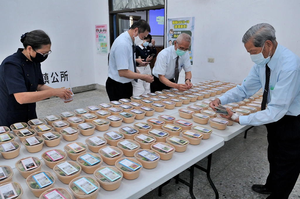 85歲的慈濟志工賴秋約(右)幫忙將兩張小卡片放在素食便當上面，讓公所同仁能夠QR CODE，上網點閱素食食譜。