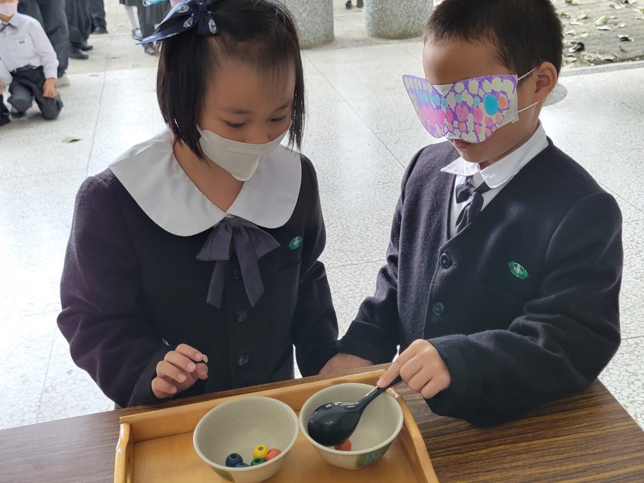 一年級小朋友戴上自製眼罩，藉由同伴的引導，用湯匙將盤中物舀至空碗中。