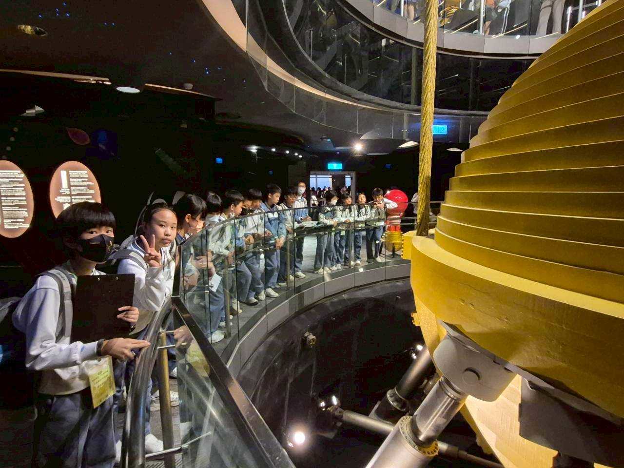 學生圍繞在台北 101 風阻尼器旁，表情驚訝或是聽導覽解說。