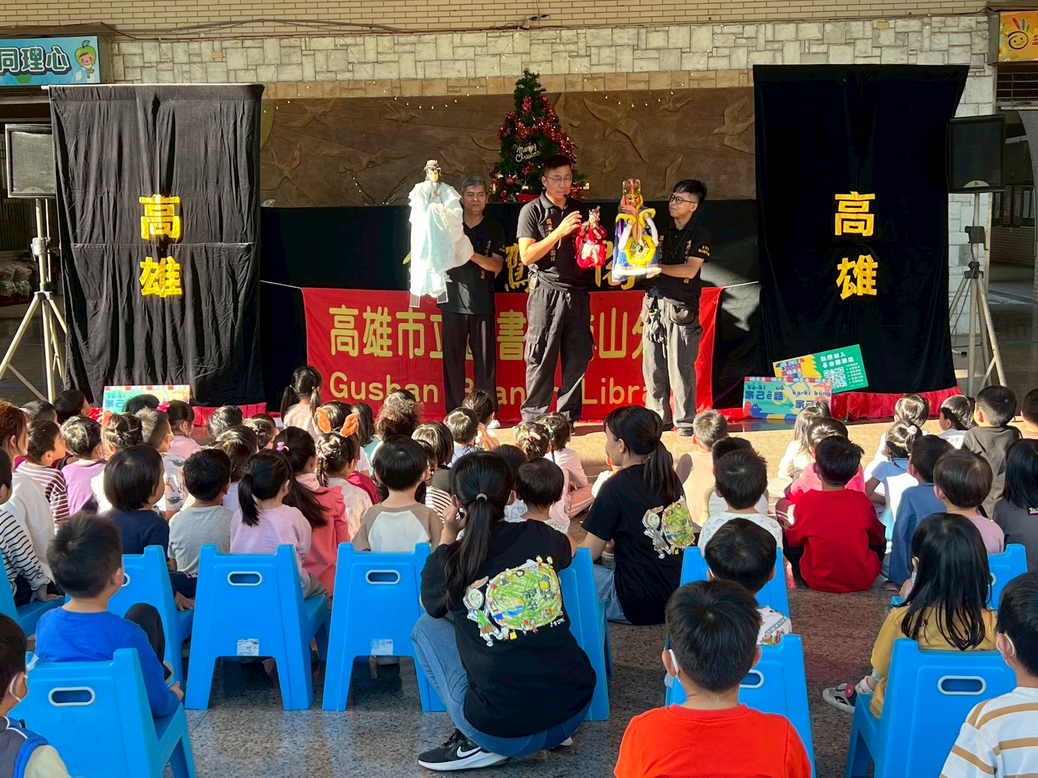 金鷹閣電視木偶劇團走進校園,操偶師在布袋戲台前向幼兒園孩子展示角色布偶,小朋友們坐在小板凳上專心觀賞,現場笑聲與掌聲不斷,氣氛熱鬧滾滾。