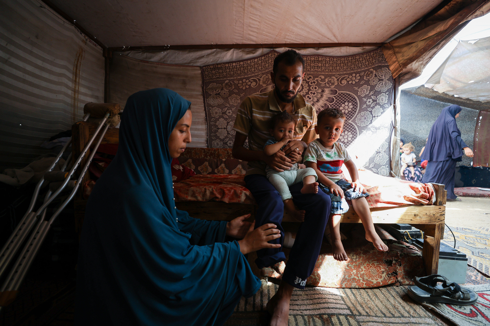 無國界醫生目睹加薩當地生活條件侵害人民基本尊嚴。Bassel一家因戰火逃離拉法（Rafah），目前住在臨時帳篷。Bassel抱著兩個孩子，妻子正在為他按摩截肢的腿，以緩解幻肢痛——數月以前，Bassel在嘗試領取物資時中彈，失去了一條腿。照片攝於2025年10月9日。