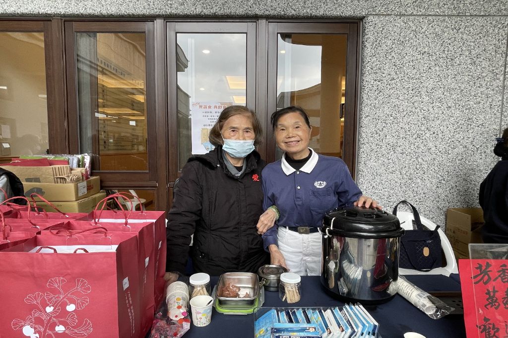 「呷菜熊蓋讚」攤位前熱鬧溫馨。年約八十歲上下的志工林鳳嬌與林謝淑美，每年歲末祝福活動總是義不容辭，熱心推廣花蓮靜思精舍師父手作的淨斯產品，與會眾廣結善緣。