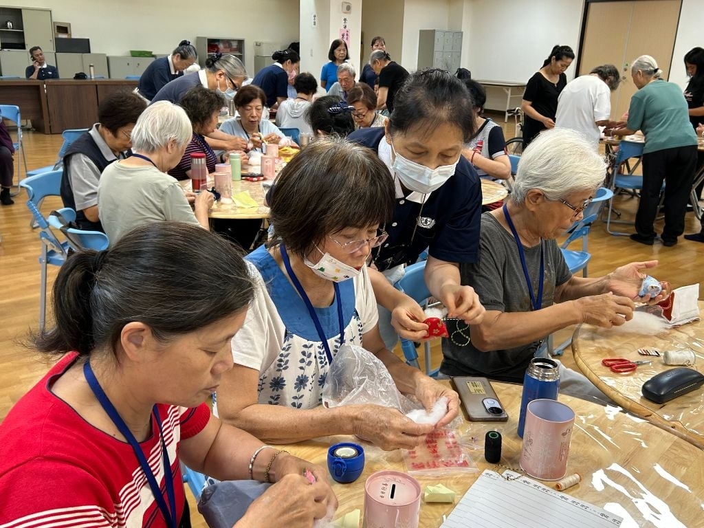 巧藝坊吳珍徵老師(中藍衣)感恩學員們，在端午佳節前夕陪伴長輩們一起動手做香包。她感性地說：「手做香包活動的氛圍，猶如兒女與長輩溫馨互動，很令人感動。這項服務不僅豐富了長者的生活，也傳遞溫暖的祝福與感恩之情。」