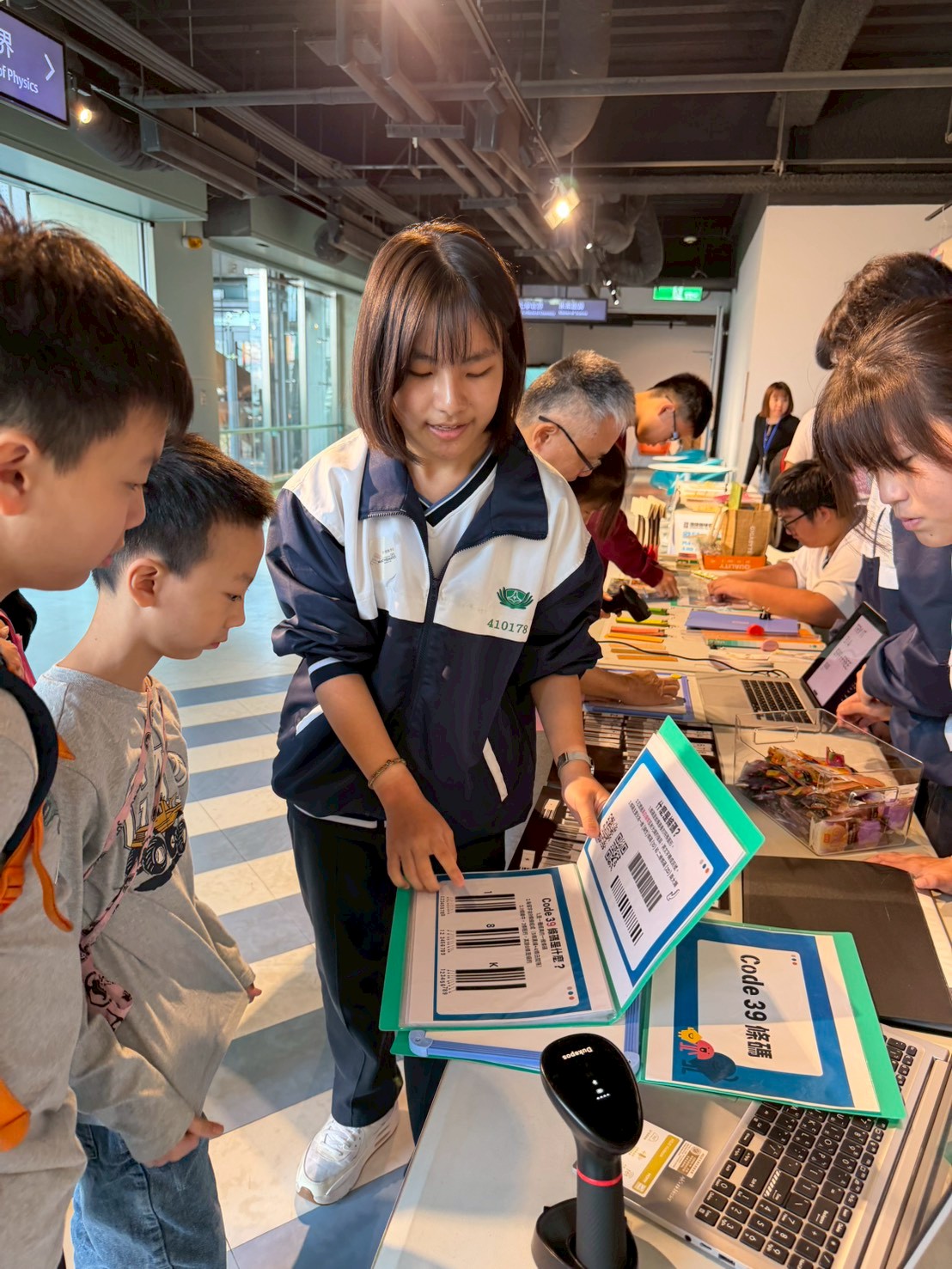 臺南慈中學生於科教館現場,利用筆電與掃碼機向家長與孩子解說 Code 39 條碼的編碼邏輯。